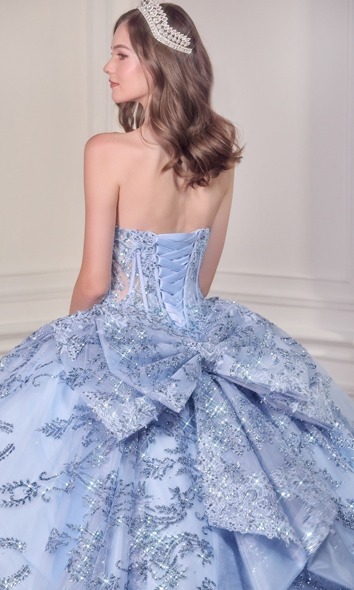 A young woman faces away from the camera, wearing the Quinceanera Dress 15732 By Ladivine—a strapless ball gown adorned with silver embellishments, lace, beaded applique, and a tiara.